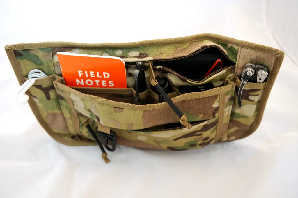 The Multicam sling bag lying open, with a notebook, pens and small tools organized in its interior pockets